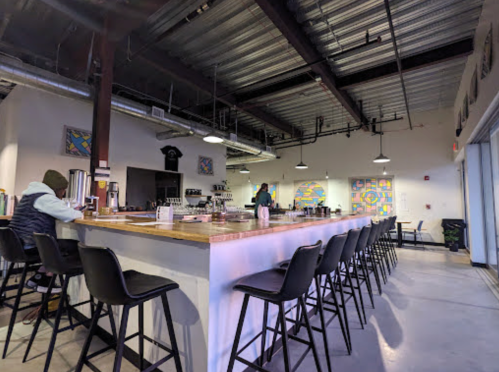 A modern café interior with a long bar, high stools, and colorful wall art. A person sits at the bar, and staff are visible.